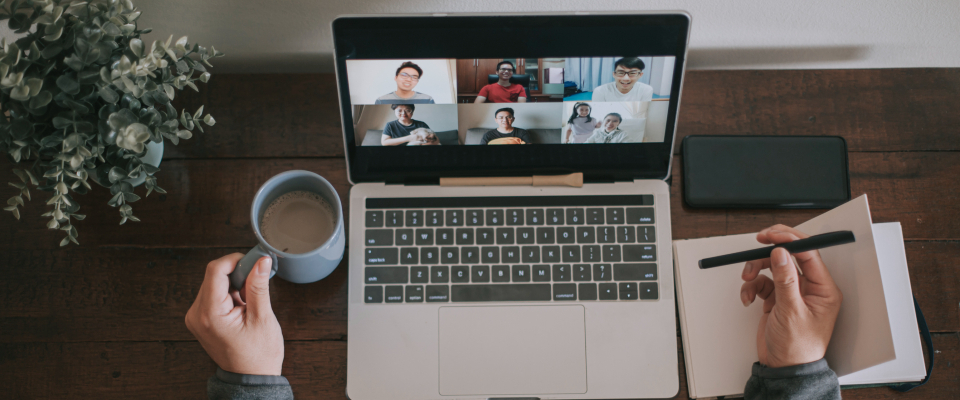 Изображение. Человек участвует в онлайн-конференции на ноутбуке, держа в одной руке чашку кофе, а в другой делая заметки в блокноте