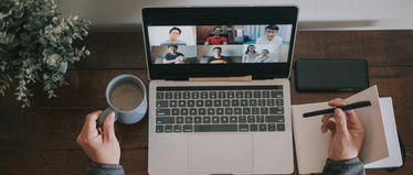 Изображение. Человек участвует в онлайн-конференции на ноутбуке, держа в одной руке чашку кофе, а в другой делая заметки в блокноте