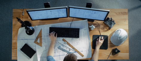 Изображение. Вид сверху на рабочее место инженера: два монитора с открытыми чертежами, клавиатура, мышь, строительные инструменты и защитная каска на столе