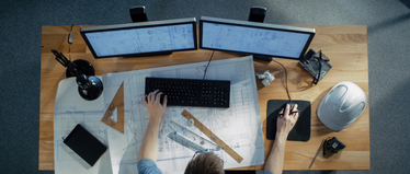 Изображение. Вид сверху на рабочее место инженера: два монитора с открытыми чертежами, клавиатура, мышь, строительные инструменты и защитная каска на столе