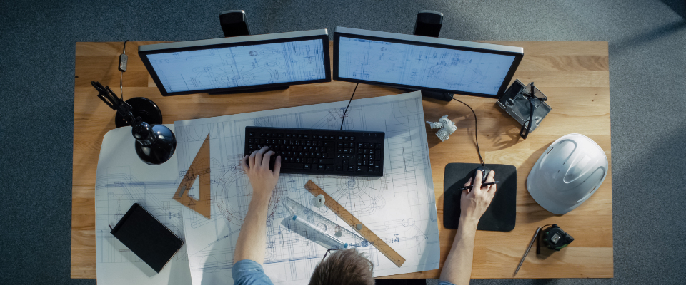 Изображение. Вид сверху на рабочее место инженера: два монитора с открытыми чертежами, клавиатура, мышь, строительные инструменты и защитная каска на столе