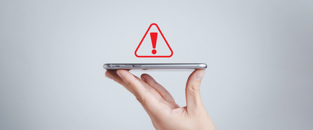 Изображение. Рука держит смартфон с красным предупреждающим треугольником и восклицательным знаком, символизирующим опасность использования мобильного устройства для бизнес-процессов