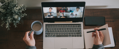 Изображение. Человек участвует в онлайн-конференции на ноутбуке, держа в одной руке чашку кофе, а в другой делая заметки в блокноте