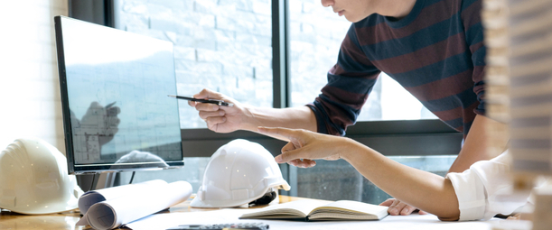 Изображение. Архитектор и дизайнер обсуждают проект. Оба указывают на монитор компьютера. Рядом лежат линейка, блокнот, чертежи, ручка и две каски.